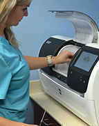 A dental professional operates a computerized milling machine in a clinic. The machine's display is active, set against a blue wall, conveying precision and focus.