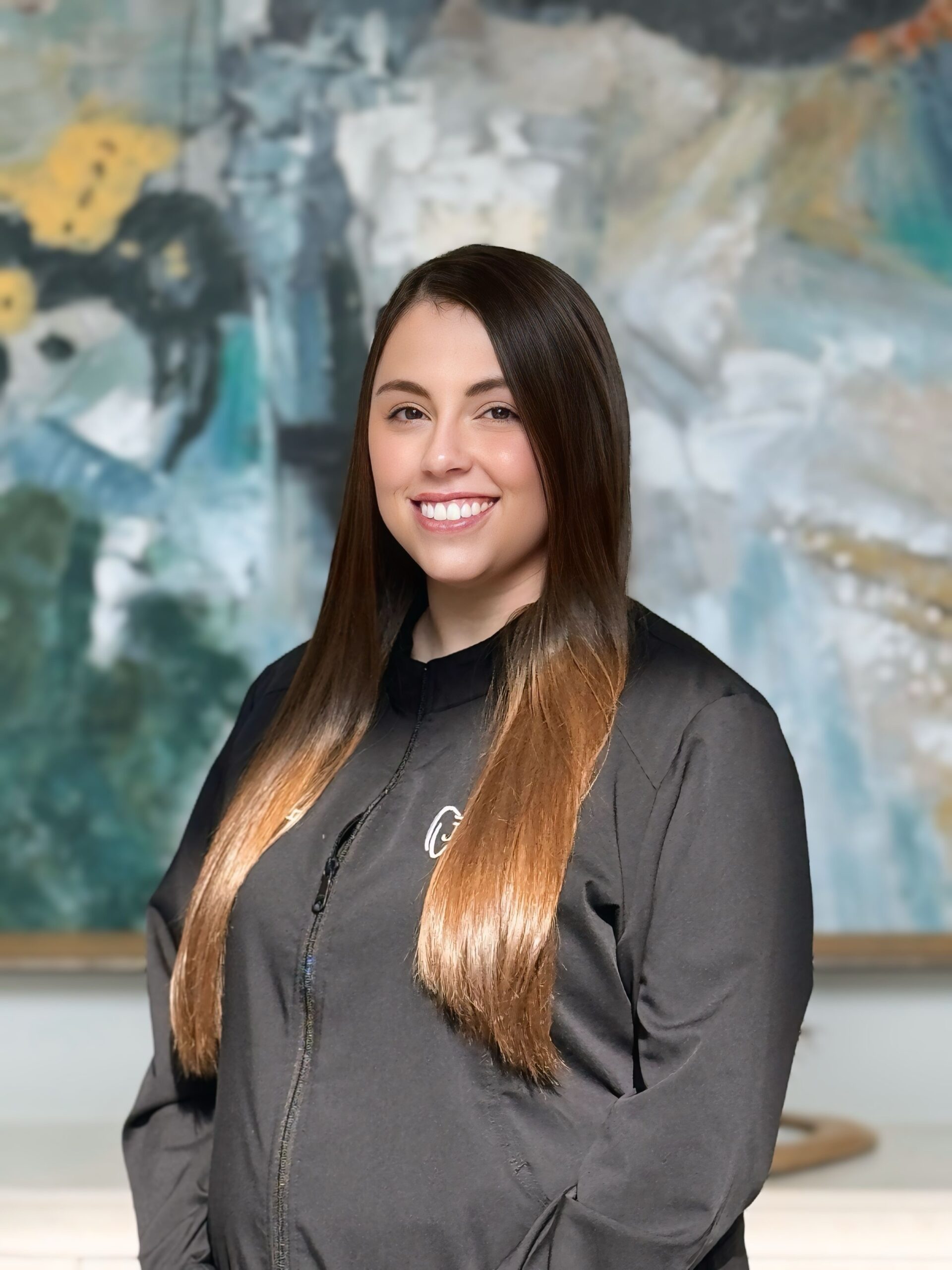 A smiling woman with long brown hair wears a black jacket, standing in front of an abstract painting with blue, green, and yellow tones. Calm atmosphere.