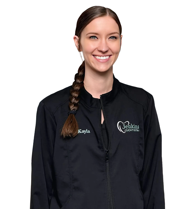 Smiling woman with a braid wears a black dental jacket with "Kayla" and "Jenkins Dentistry" logos. Abstract art hangs in the blurred background.