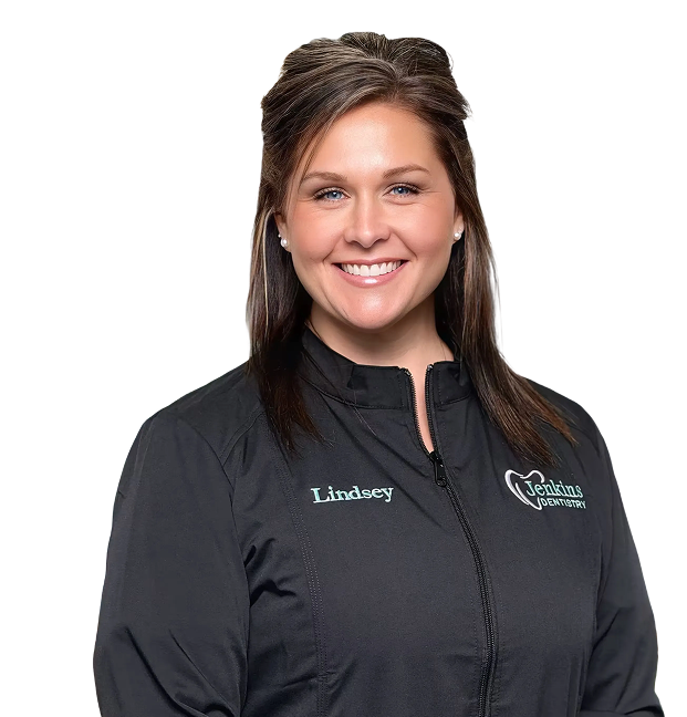 A woman with brunette hair smiles warmly, wearing a black jacket with "Lindsey" embroidered on it.