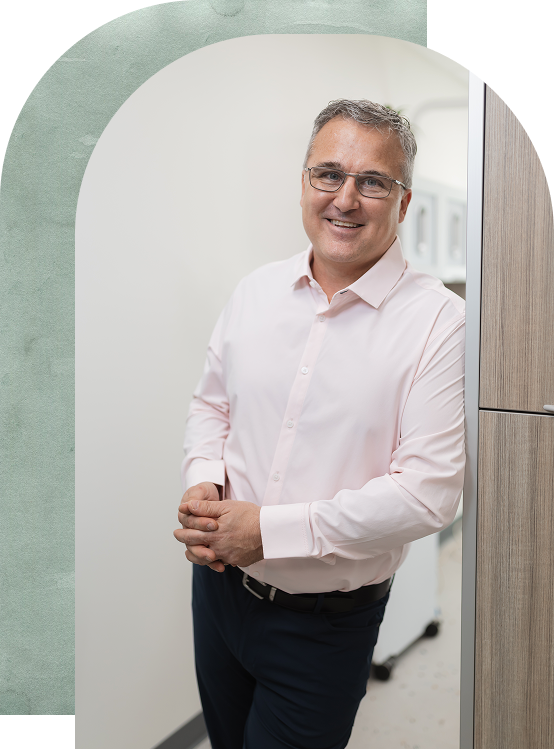 Smiling man in a light pink shirt leans casually against a wall in a modern office hallway. The scene conveys a professional and approachable demeanor.