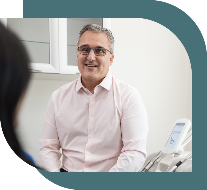 Man in glasses and a light pink shirt smiles warmly, seated in a bright medical office. Medical device visible, suggesting a professional setting.