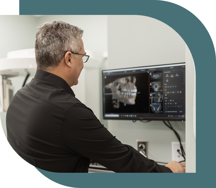 A man in a black shirt views dental X-rays on a computer monitor in a modern clinic. The scene suggests a professional, focused atmosphere.
