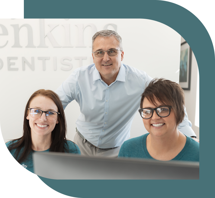 A smiling dentist and two dental assistants, all wearing glasses, sit in a modern dental office. The atmosphere is friendly and professional.