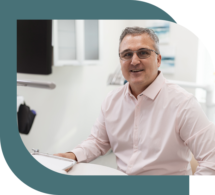 A smiling man with glasses and a pink shirt sits at a desk in a bright office, conveying a welcoming and professional atmosphere.