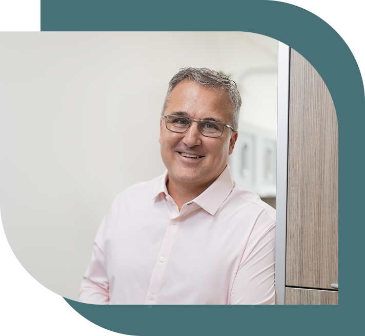 Smiling man with glasses and salt-and-pepper hair, wearing a light pink shirt, stands in a bright, modern space with wooden cabinets. Confident and approachable.