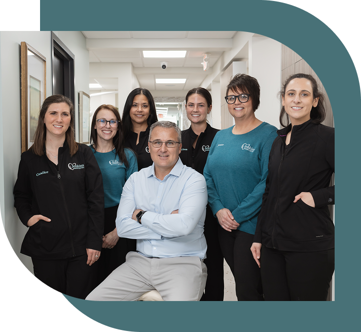 A group of seven smiling people wearing professional attire in a well-lit hallway. The central figure, seated, is surrounded by six standing colleagues.