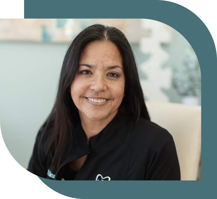 Smiling woman with long dark hair in a black jacket sits indoors against a blurred light background, conveying warmth and approachability.
