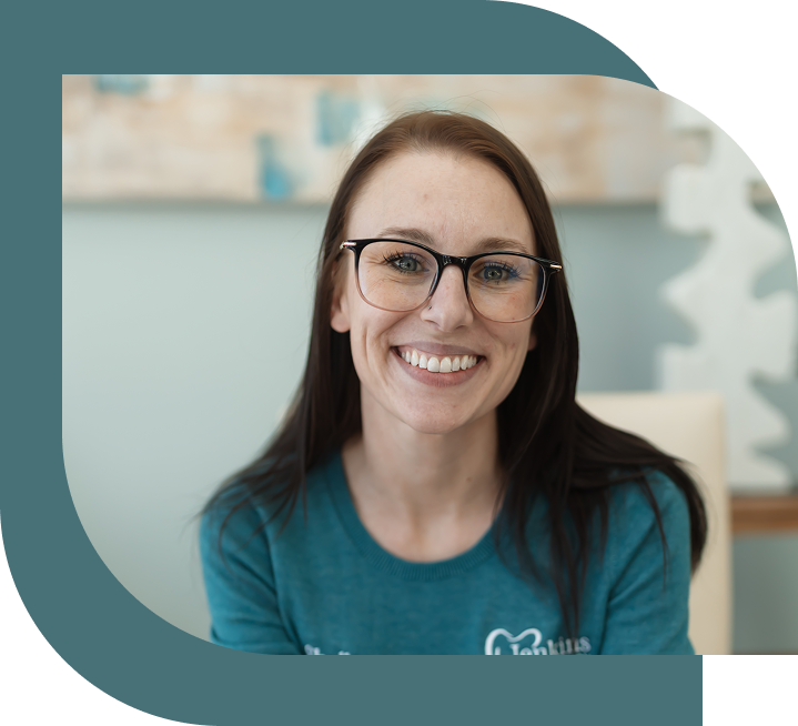 Smiling woman with brown hair and glasses wearing a turquoise sweater. She is in a softly lit room with abstract art in the background, conveying warmth.