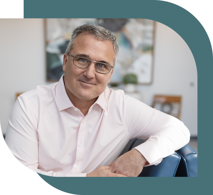 Middle-aged man with glasses, wearing a light pink shirt, smiles softly while seated in an office. Background features a blurred abstract painting.