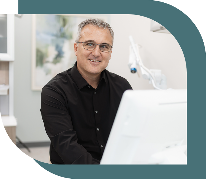 A person wearing glasses and a black shirt smiles confidently at a desk in a modern, well-lit office, conveying a welcoming and professional tone.