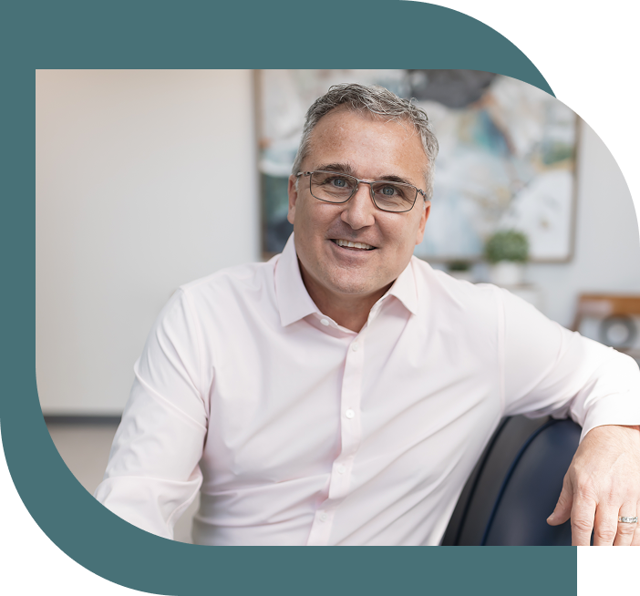 A smiling man in a white shirt and glasses leans on a chair in a modern office with abstract art and plants, creating a welcoming, professional tone.