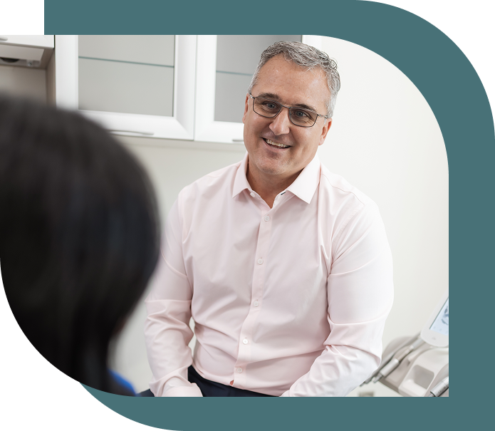 A smiling man in glasses and a light pink shirt sits in an office, engaging with someone off-frame. The setting is warm and professional.