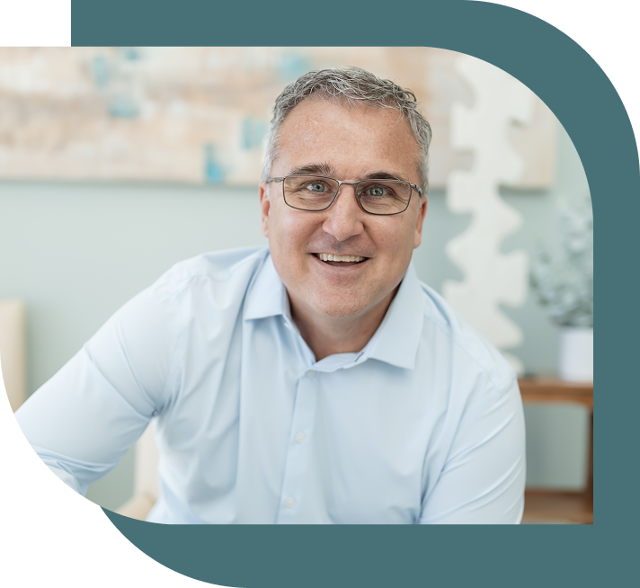 A smiling man with glasses and graying hair wears a light blue shirt, sitting in a softly lit modern room with blurred abstract art in the background.