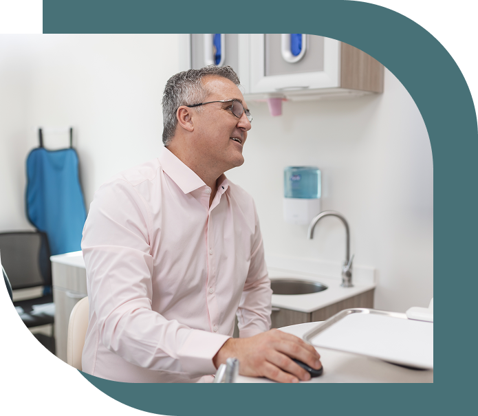 A man wearing glasses and a light pink shirt smiles while sitting in a modern office with a sink and medical equipment in the background.