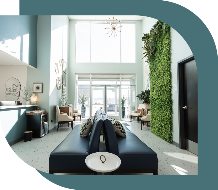 Bright, modern dental office waiting area with high ceilings, large windows, and a green plant wall. Blue seating, decorative pillows, and stylish lighting.