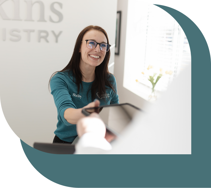 A woman with long brown hair and glasses, wearing a teal shirt with "Shelby" on it, smiles while handing a clipboard in a bright reception area.