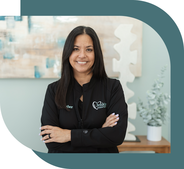 Smiling woman with long dark hair, in a black jacket with "Jenkins Dentistry" logo, against a black background, arms crossed confidently.