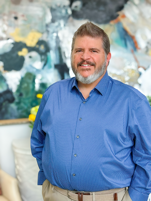 Smiling man in a blue shirt and beige pants stands with hands in pockets. The background is a blurred colorful mural, creating a cheerful atmosphere.