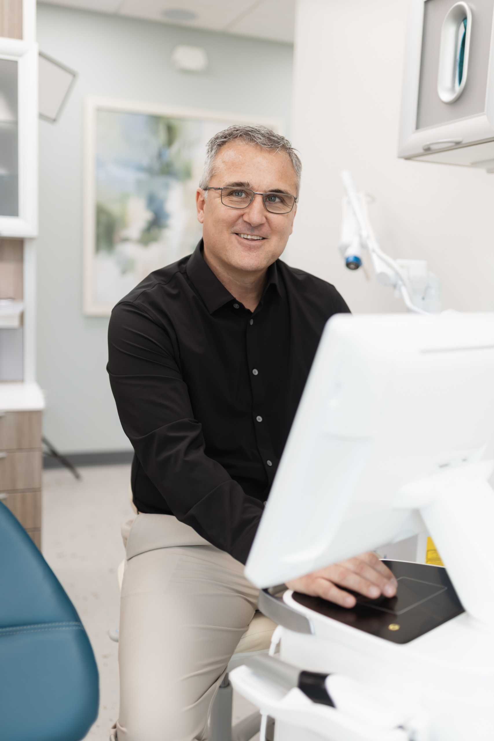 A man in business casual attire sits at a desk in a modern office, smiling at the camera with one hand on a computer keyboard.
