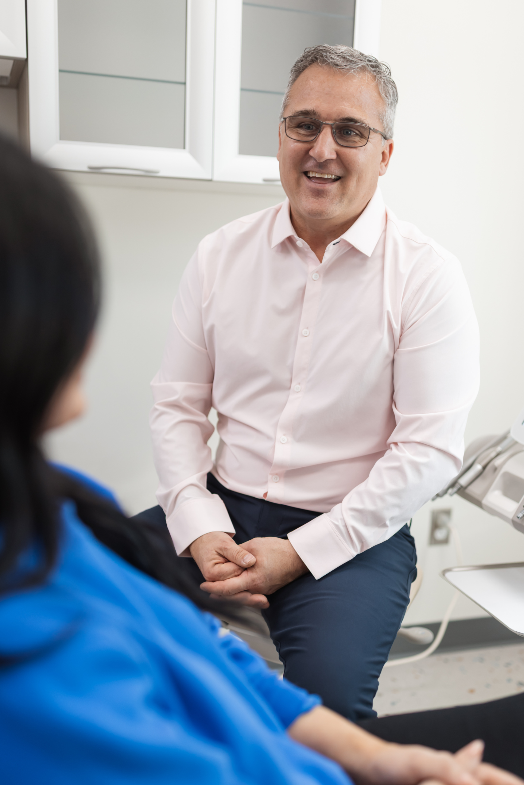 A man in a light pink shirt sits in a medical office, facing and speaking with a seated person wearing a blue top. Medical equipment is visible nearby.
