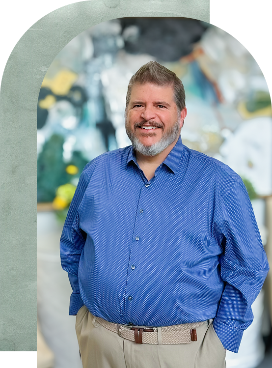 Smiling man in a blue shirt and beige pants stands with hands in pockets. The background is a blurred colorful mural, creating a cheerful atmosphere.