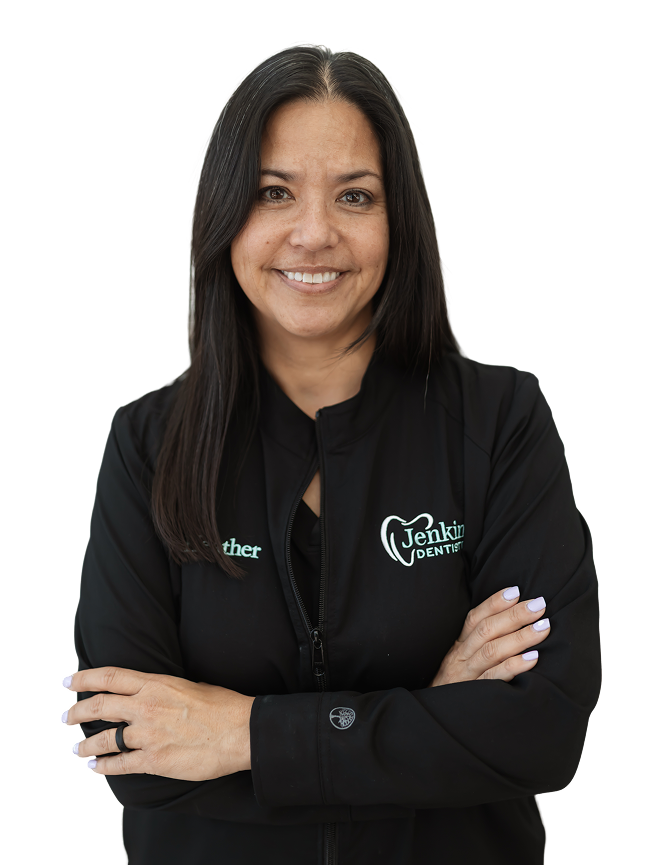 Smiling woman with long dark hair, in a black jacket with "Jenkins Dentistry" logo, against a black background, arms crossed confidently.